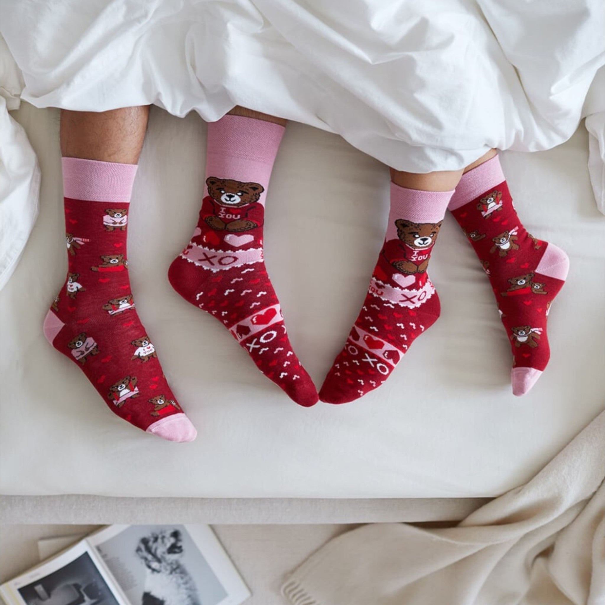 Vrolijke rode sokken met teddyberen en hartjes van Many Mornings, gedragen in bed onder een wit dekbed.