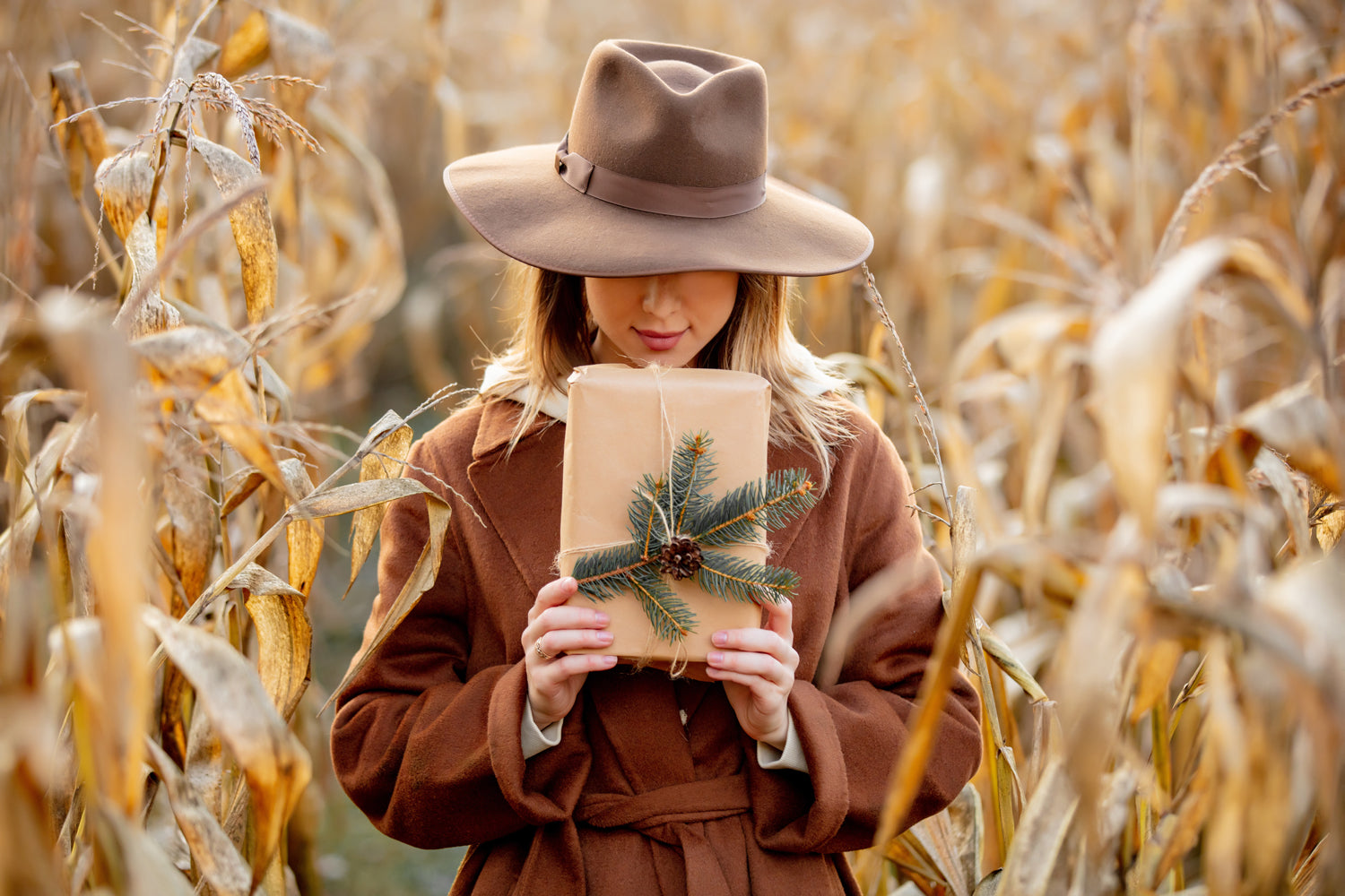Vrouw in bruine herfstjas en hoed houdt cadeau met dennentak vast tussen verdorde maïsstengels, sfeerbeeld van de herfstcampagne van Omdat.
