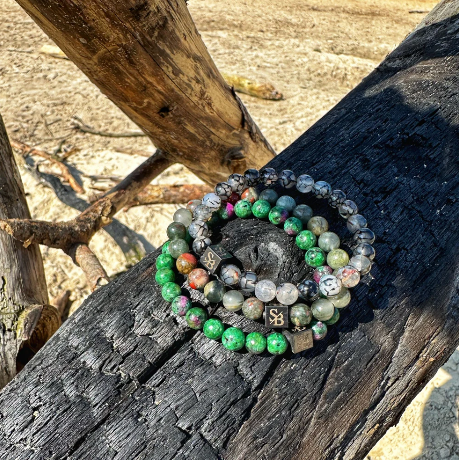 Meerdere Stoney Bracelets armbanden, African Bloodstones, Dragon Veins en Aqua Terra Stones, gepresenteerd op zwartgeblakerde houtstam in het zand.