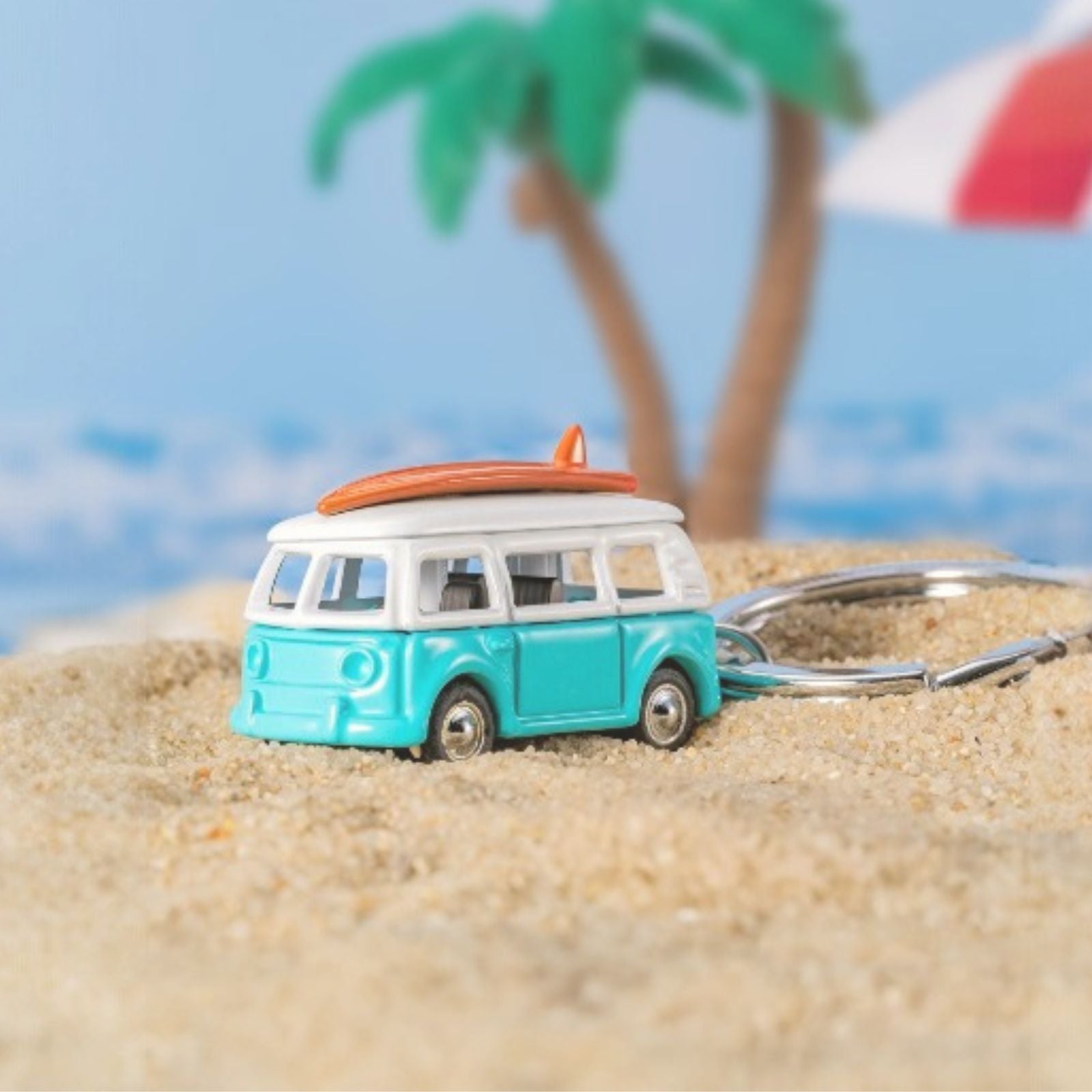 Sleutelhanger van Metalmorphose in de vorm van een turquoise surfbusje, gefotografeerd in zomerse strandsfeer.