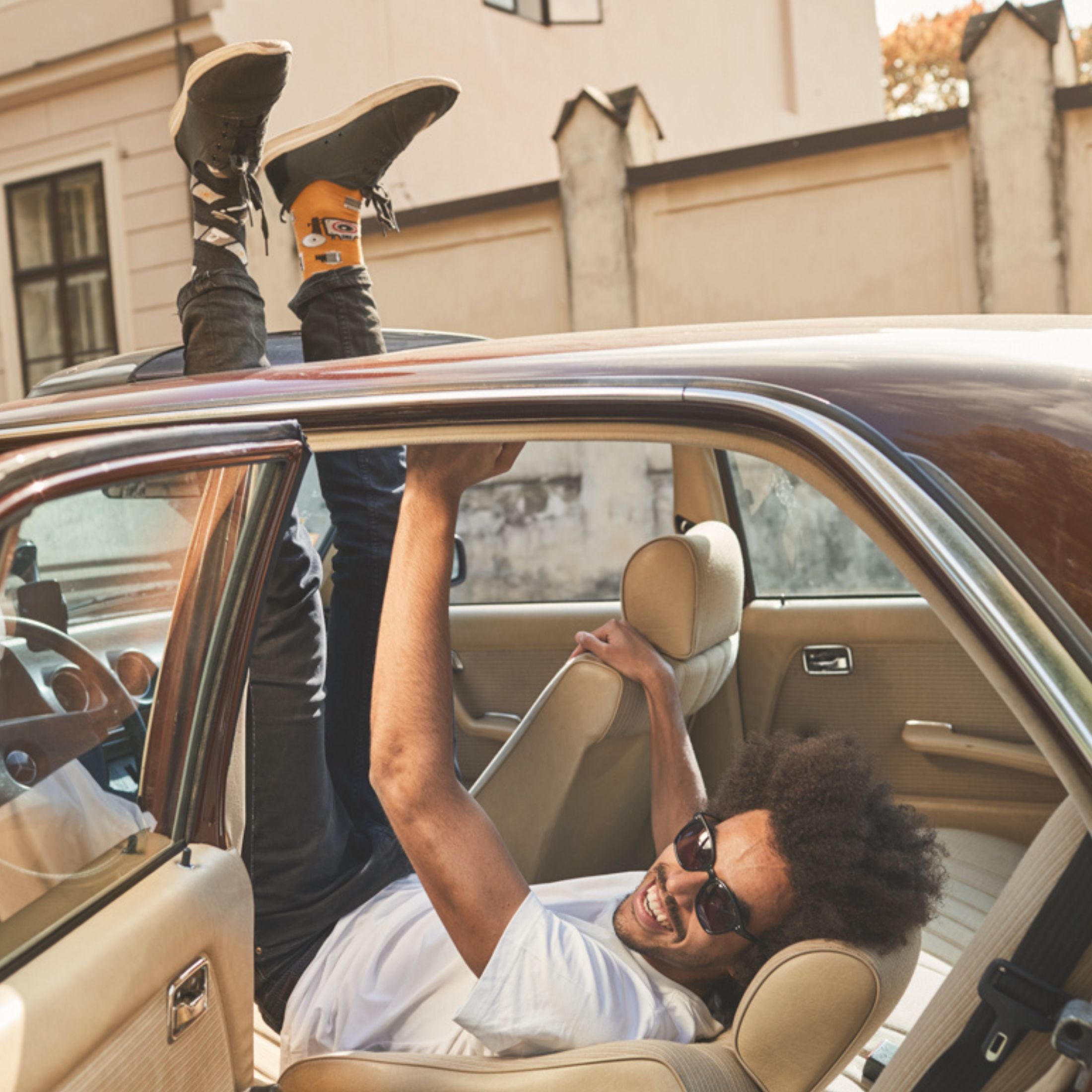 Lachende man in auto met benen omhoog, draagt opvallende sokken van Many Mornings met geel en zwart design.
