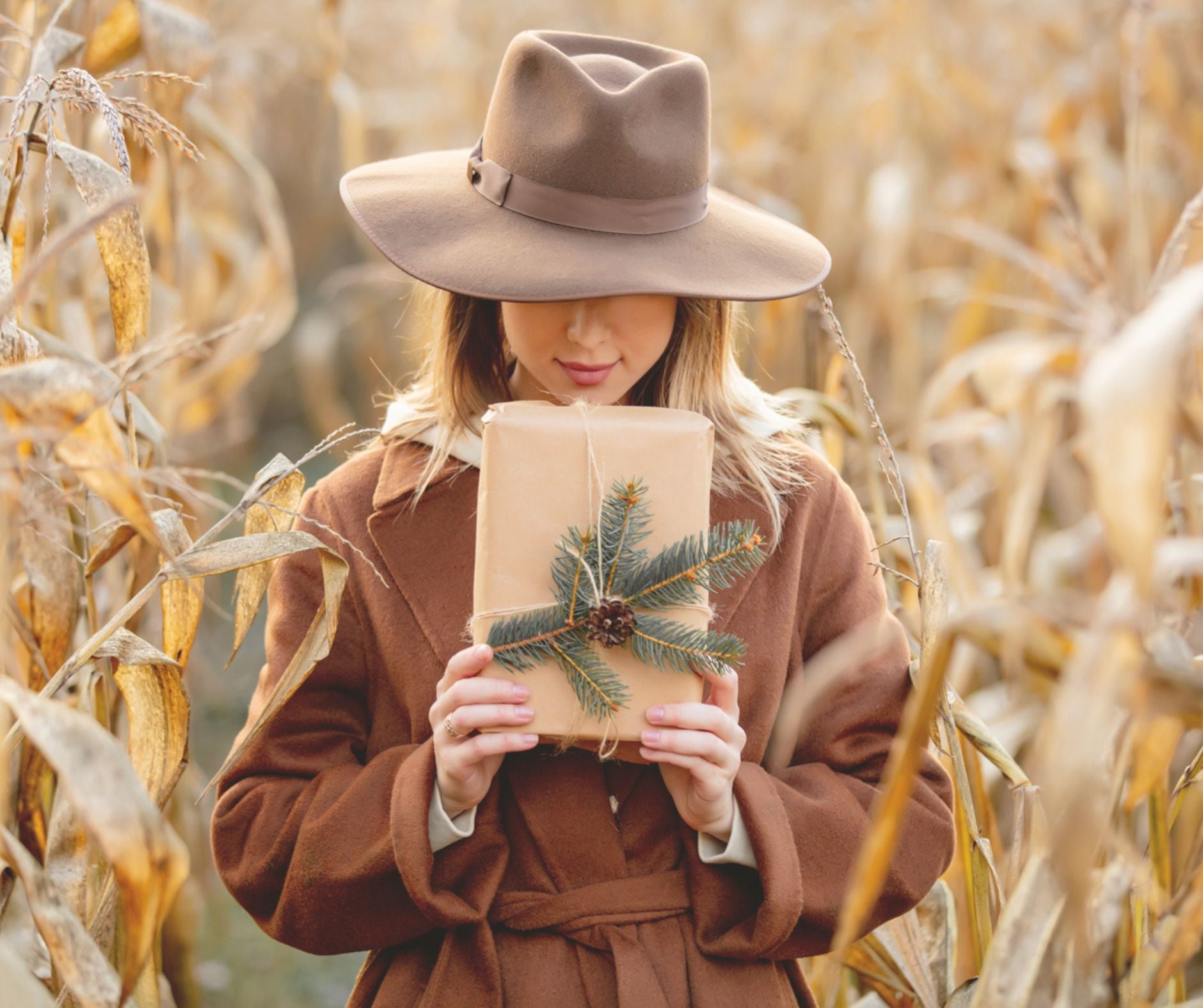 Vrouw in bruine herfstjas en hoed houdt cadeau met dennentak vast tussen verdorde maïsstengels, sfeerbeeld van de herfstcampagne van Omdat.