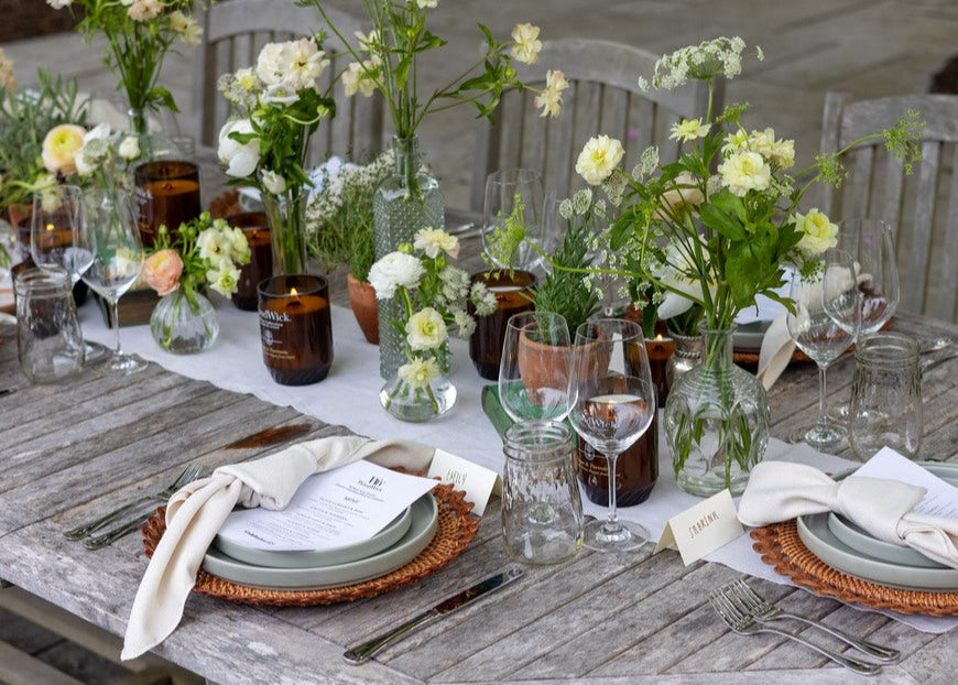 Sfeervol gedekte buitentafel met WoodWick Renew sojawaskaarsen in bruine glazen potten, omringd door bloemen en wijnglazen, verkrijgbaar bij cadeauwinkel Omdat in Uden.