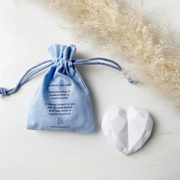 Witte hartvormige bruisbal van Rockstyle met edelsteen, gepresenteerd naast een blauw linnen zakje met tekst, op een lichte ondergrond met decoratief pampasgras.