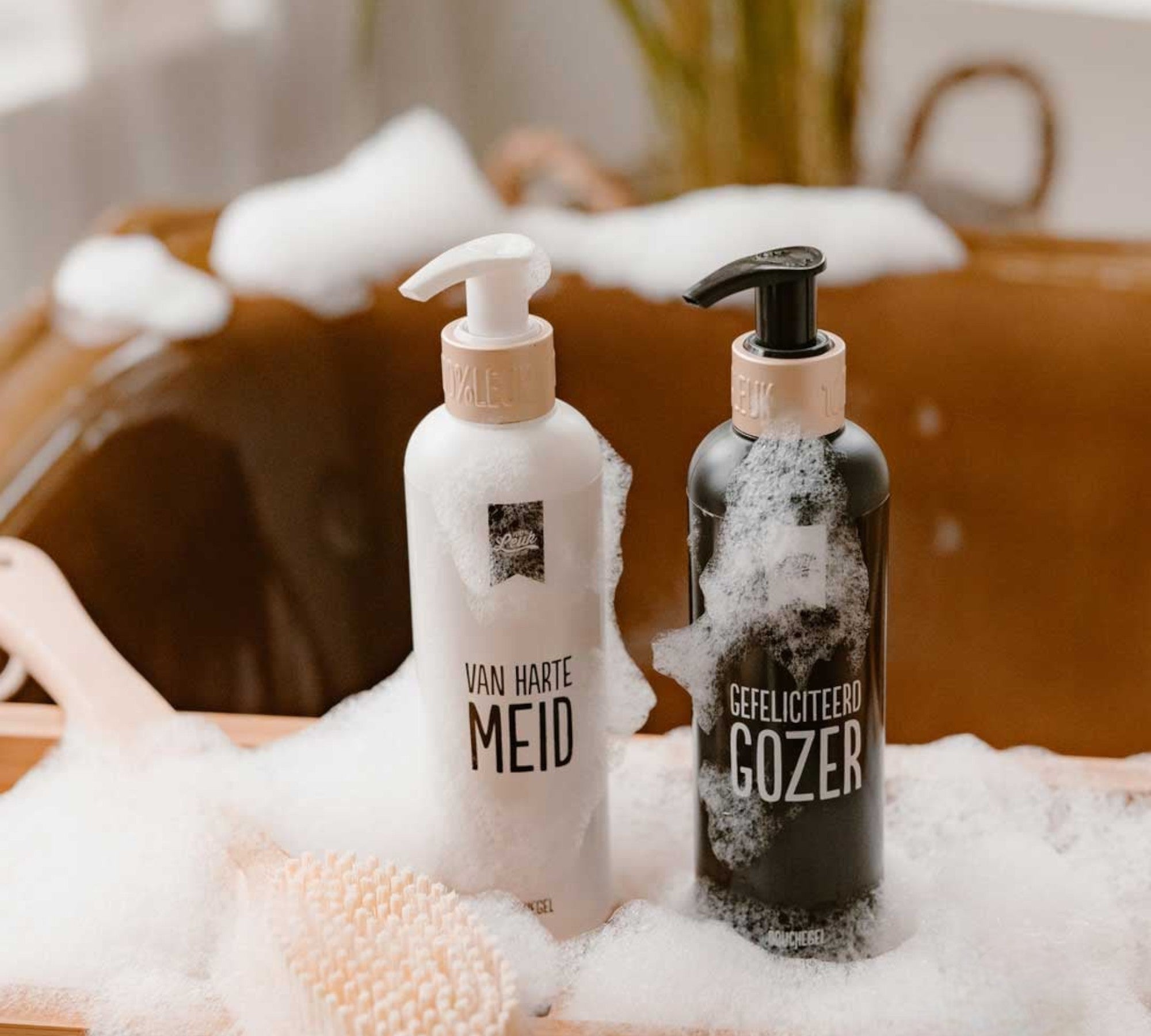 Twee douchegels van 100%Leuk met grappige teksten "Van harte meid" en "Gefeliciteerd gozer", sfeervol gefotografeerd in een schuimbad met houten borstel.