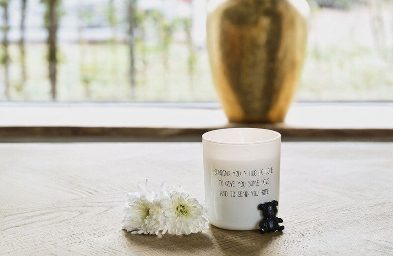 Witte geurkaars met de tekst ‘Sending you a hug to give you some love and to send you hope’ – troostkaars met bloemen en een klein zwart beertje ernaast.